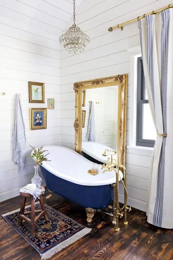 a refined bathroom with white shiplap, a navy clawfoot bathtub, a floor mirror in a gold frame, a crystal chandelier and artworks