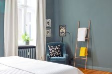 19 a stylish bedroom with slate blue walls, a bed, a corner with a blue chair, black and white artwork, a ladder and some blank space for an airy feel