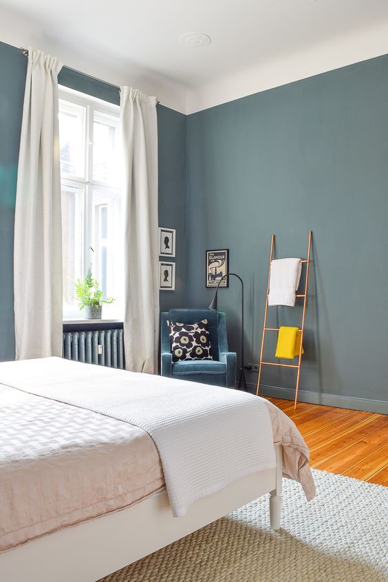 a stylish bedroom with slate blue walls, a bed, a corner with a blue chair, black and white artwork, a ladder and some blank space for an airy feel