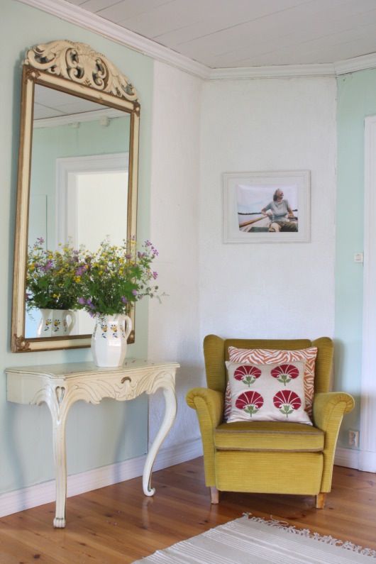 a vintage nook with a white carved half console table, a mirror in a vintage frame, a mustard chair with a bright pillow and some blooms