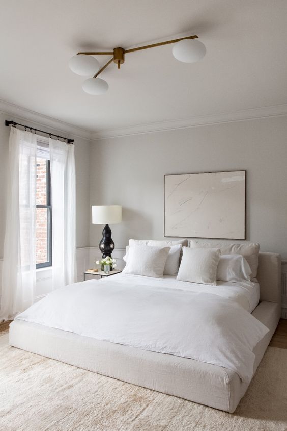 a neutral minimalist bedroom with paneled walls, an upholstered bed, neutral bedding, a neutral artwork, table lamps and nightstands, some negative space