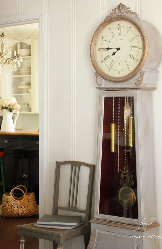 a vintage cottage space with a whitewashed grandfather clock, a grey chair, a black kitchen island with butcherblock countertop