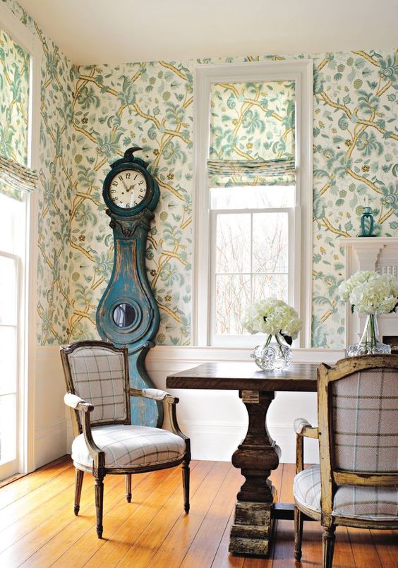 a vintage dining space with botanical wallpaper, a rich-stained dining table, neutral chairs, a navy shabby chic grandfather clock and blooms