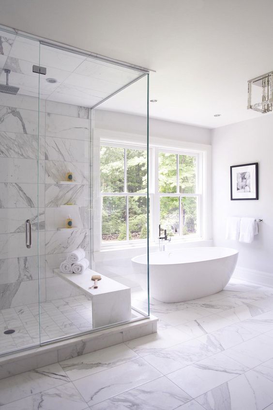 a neutral bathroom clad with white marble tiles, an oval tub by the window, a shower space and blank space for a more luxurious look