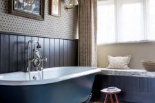 26 a vintage bathroom with printed wallpaper, navy paneling, a navy clawfoot bathtub, a windowsill bench and printed textiles is ultimately chic