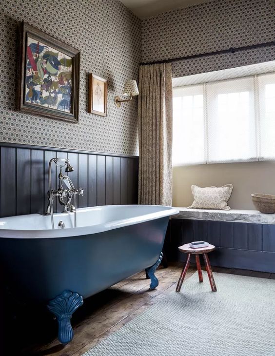 a vintage bathroom with printed wallpaper, navy paneling, a navy clawfoot bathtub, a windowsill bench and printed textiles is ultimately chic