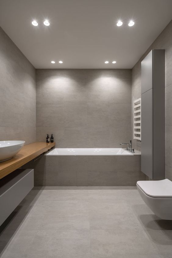 a minimalist bathroom clad with grey large scale tiles, with a floating shelf vanity, a bathtub clad with tiles and a toilet