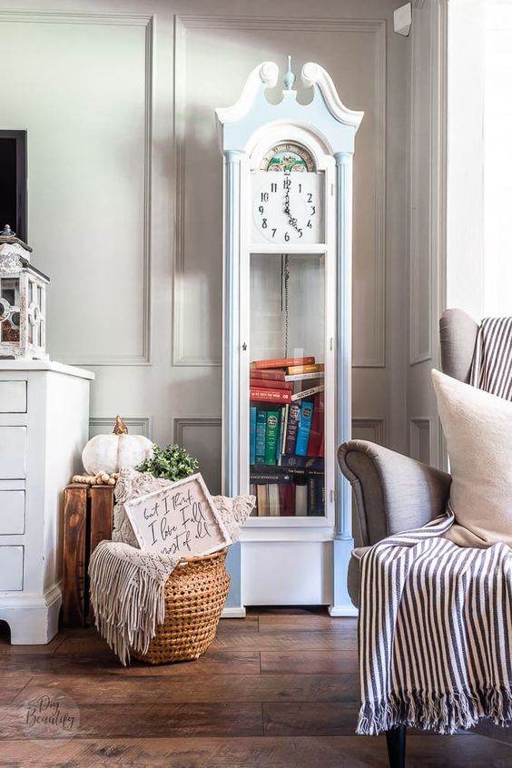 a vintage farmhouse space with a blue and white grandfather clock, a grey chair with textiles, a basket with blankets and greenery
