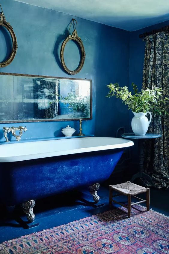 a vintage inspired bathroom with blue walls and an electric blue bathtub, a bold printed rug, a vintage mirror, greenery and printed curtains
