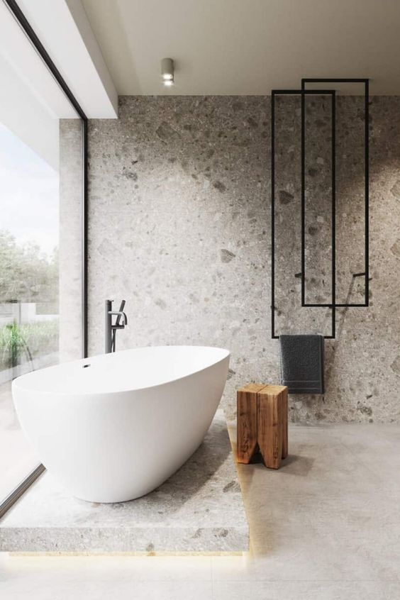 a minimalist bathroom clad with grey terrazzo, with a platform and a bathtub on top, black radiators and a glazed wall