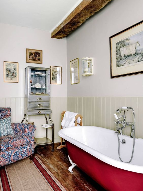 an English country bathroom with neutral wall paneling, a bright printed chair, a burgundy clawfoot bathtub, a wooden beam and some art