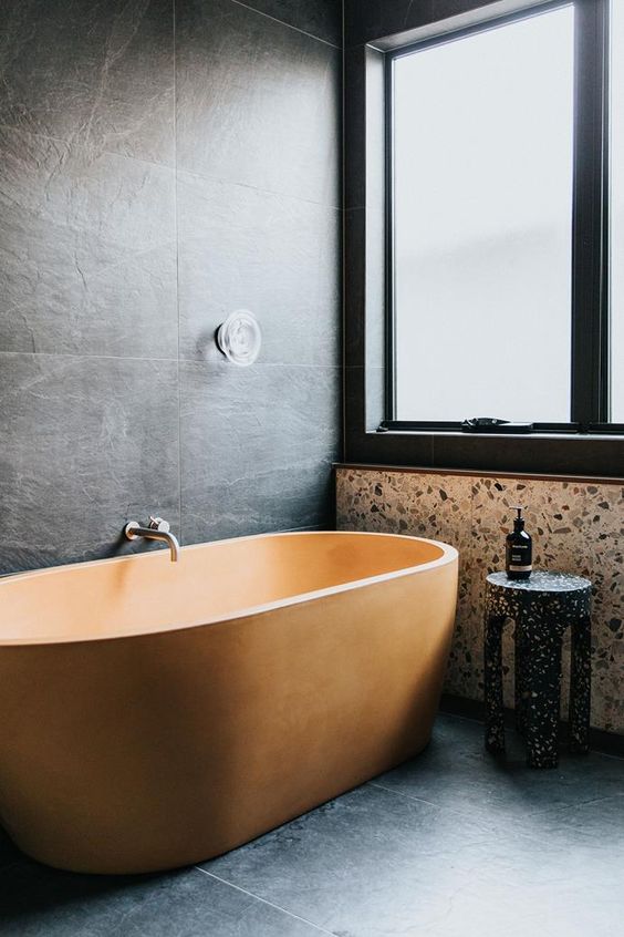 a moody minimalist bathroom clad with large scale tiles, terrazzo in neutrals and black, a beautiful and textural orange bathtub