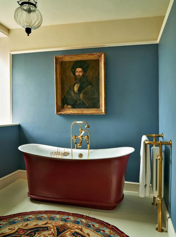 a vintage bathroom with blue walls, a modern deep red bathtub, gold fixtures and a vintage artwork plus a colorful printed rug just wows