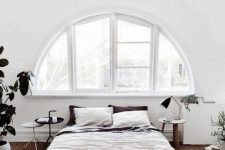 35 an airy white Nordic bedroom with an arched window, a bed, some wooden furniture and potted greenery