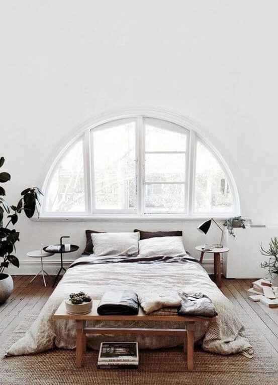 an airy white Nordic bedroom with an arched window, a bed, some wooden furniture and potted greenery
