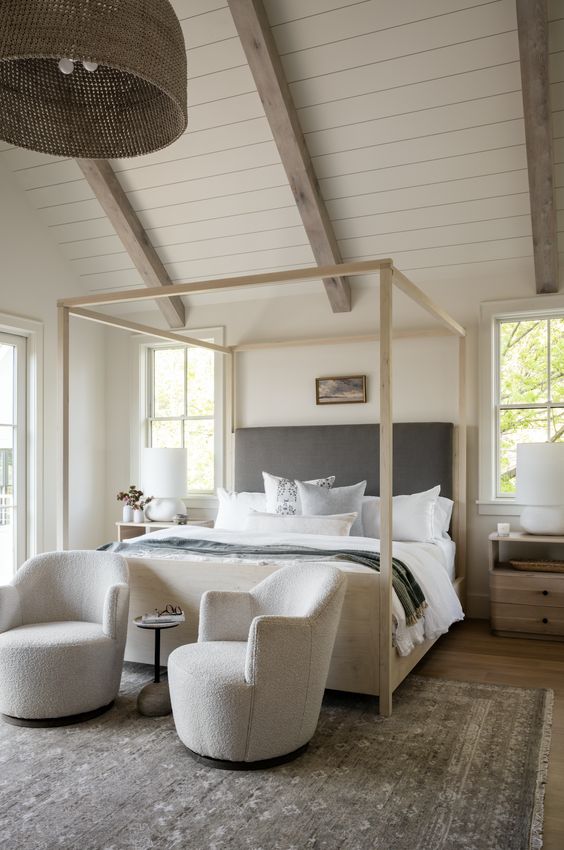 a beautiful and cozy neutral bedroom with shiplap on the ceiling, a frame bed with neutral bedding, light-stained nightstands and creamy chairs at the foot of the bed
