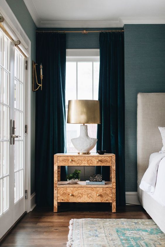a beautiful bedroom with blue grasscloth wallpaper, teal velvet curtains, a neutral upholstered bed, a light-colored nightstand and a gold lamp