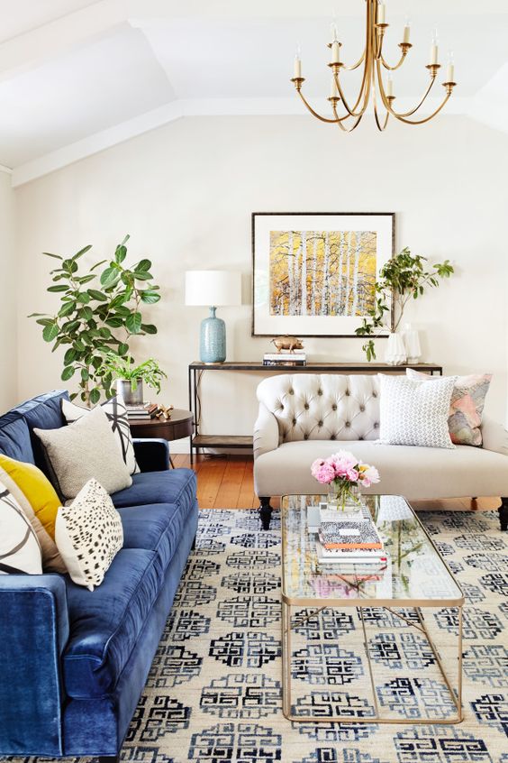 a beautiful living room with a printed rug, a blue sofa, neutral and printed pillows, a creamy sofa, greenery and a gold chandelier