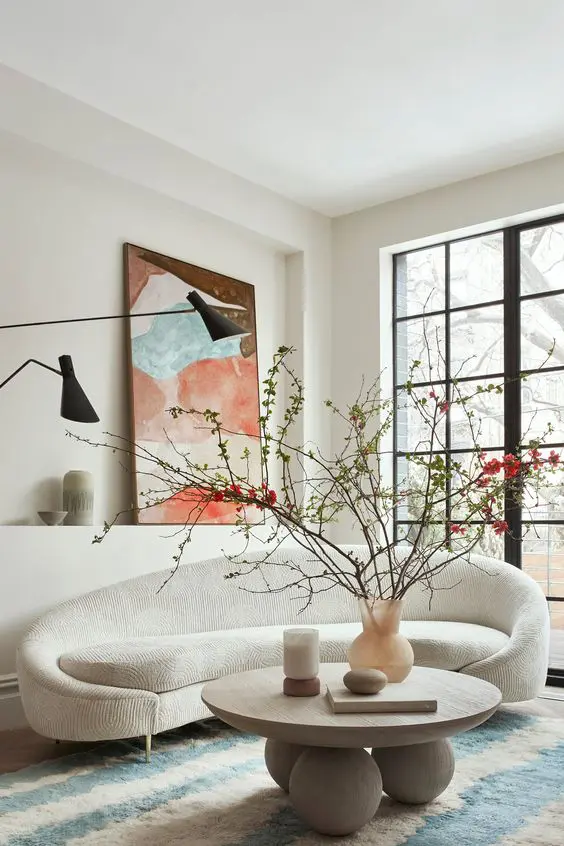 a beautiful small living room with a curved creamy sofa, a wooden coffee table, a bright artwork, black lamps and a glazed wall