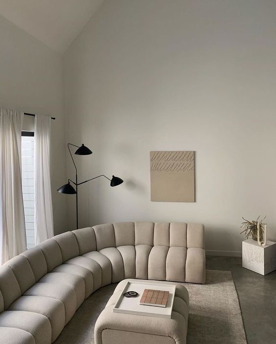 a beautiful warm colored neutral living room with a creamy curved sofa and a matching pouf, a black floor lamp and a cube