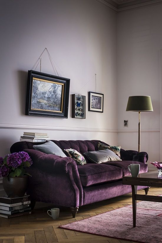 a blush living room with a refined purple sofa, a dusty pink rug, a coffee table, a floor lamp, a gallery wall and a stack of books