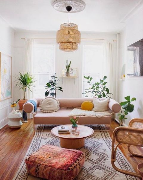 a boho living room with a blush sofa, a printed rug and a pouf, a round table, potted plants and a cane chair plus a woven pendant lamp