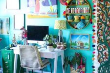 a boho maximalist home office with a turquoise accent wall, a white desk, a green cabinet and lots of artworks and textiles in bold shades