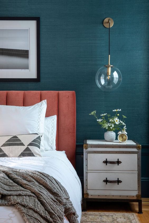 a bold bedroom with teal grasscloth wallpaper, a coral bed with pretty bedding, a sconce and a white nightstand with blooms