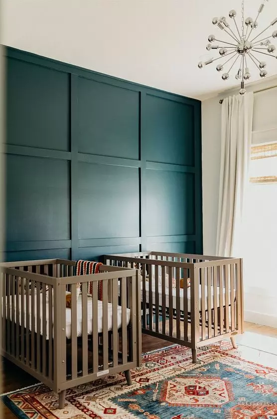 a bold twin nursery with a navy paneled wall, neutral cribs, a disco ball chandelier and a bright printed rug