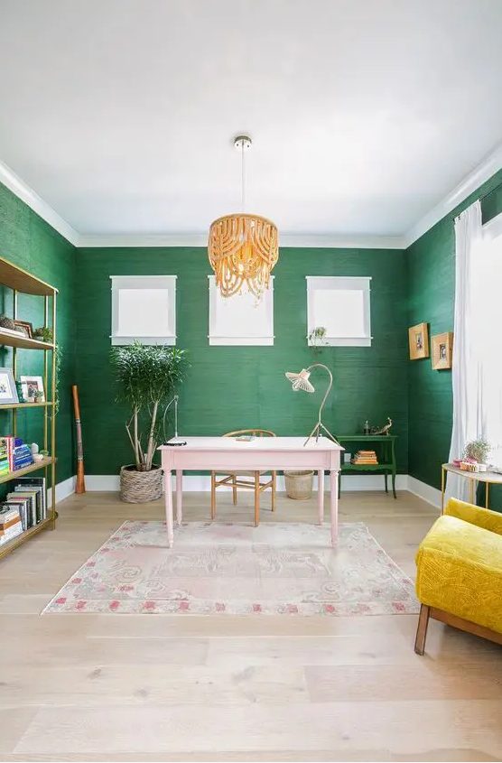 a bright home office with green grasscloth wallpaper, a pink desk, a yellow chair, a beaded chandelier and a brass storage unit