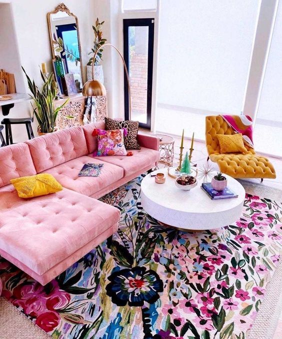 a bright living room with a pink sectional, a bold floral rug, a round coffee table, a mustard tufted chair, bright pillows and decor