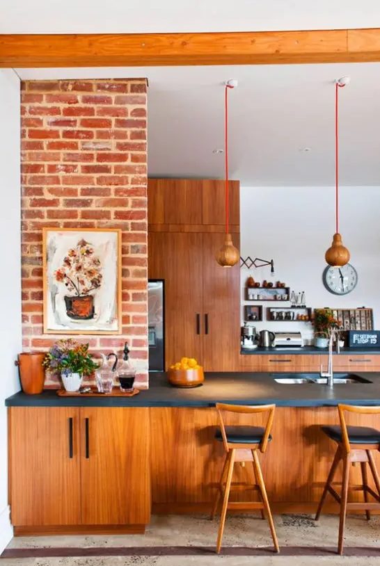 a bright mid-century modern kitchen with reddish furniture, pendant lamps, black countertops and artworks