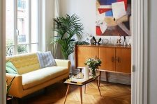 a bright modern living room with a light yellow sofa, a colorful artwork, mid-century modern furniture and greenery