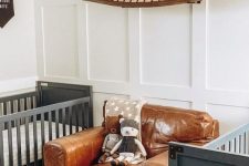 a catchy adventurous twin nursery with grey cribs, a leather chair, a canoe, an antler chandelier and white paneled walls