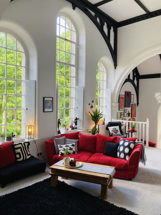 a catchy living room with double-height arched windows, a red sofa with printed pillows and a blakc low seat sofa, a wooden table and potted greenery