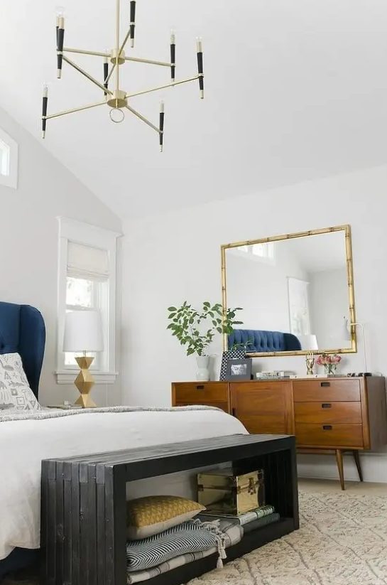 a chic and bright mid century modern bedroom with white walls, rich stained furniture, a black storage bench and a gilded chandelier
