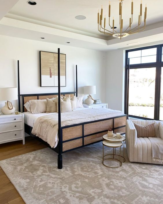 a chic and refined neutral bedroom with a black wood and cane bed, a neutral chair, a printed rug, a gold chandelier and white nightstands