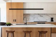 a chic contemporary kitchen with white and light stained cabinets, a large kitchen island with a white stone countertop and a backsplash, a black pendant lamp