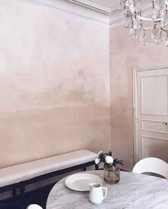 a chic dining room with blush limewashed walls, a stone tabletop dining table, an upholstered bench, a crystal chandelier