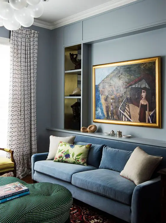 a chic living room done in light blue, with a blue sofa with pillows, niches for storage, a green ottoman and printed curtains