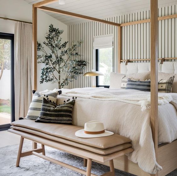 a chic neutral bedding with striped wallpaper, a light stained bed with neutral bedding, a leather and wooden bench and a potted plant