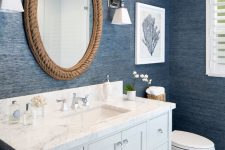 a coastal bathroom with navy grasscloth wallpaper, a white vanity with a stone countertop, white appliances and a mirror in a rope frame