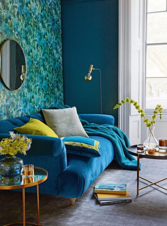 a colorful living room with a bold printed accent wall, a bright blue sofa with pillows and a blanket, round tables and floor lamps