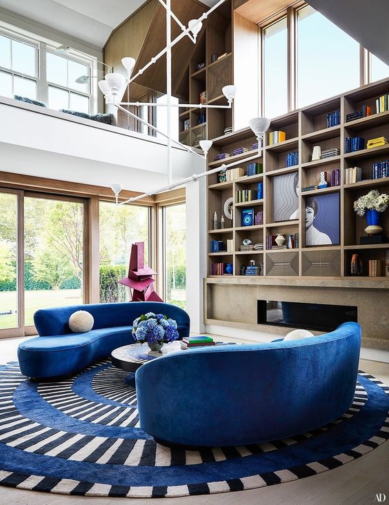 a colorful living room with a built in fireplace, a large built in bookcase, bright blue curved sofas, a striped rug and a coffee table