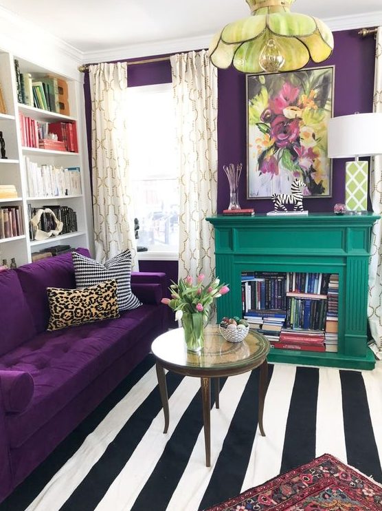 a colorful living room with a deep purple accent wall and a matching sofa, a striped rug, a floral pendant lamp and an emerald fireplace with books