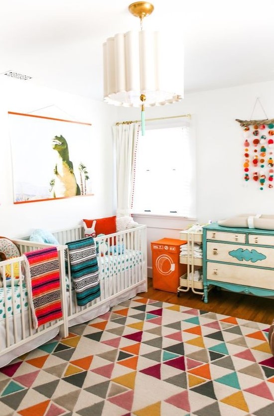 a colorful mid-century modern nursery with white cribs, a neutral and turquoise dresser, colorful bedding and a colorful rug plus a cool chandelier