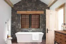 a contemporary bathroom with a stained wooden floor, a stone accent wall, a tub and a small side table, wooden beams and a wooden vanity