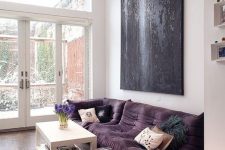 a contemporary living room done in neutrals, with a deep purple sofa and an ottoman, a statement black artwork and a white coffee table