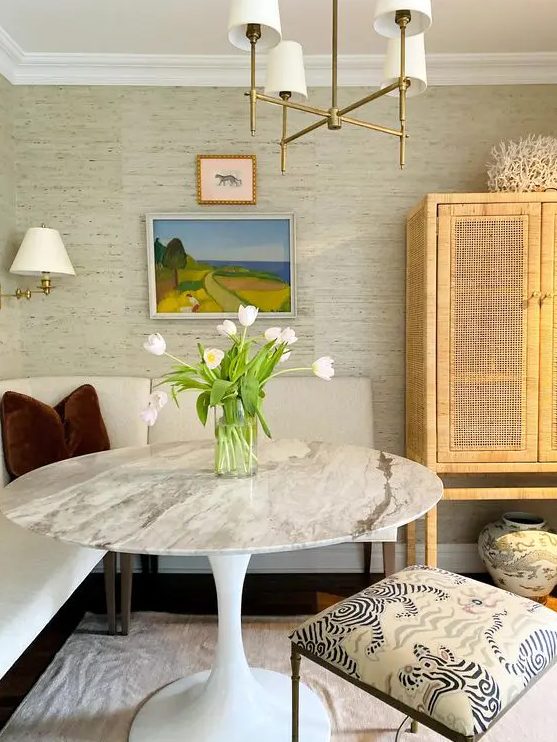 a cozy dining nook with neutral grasscloth wallpaper, a creamy banquette seating, a round marble table and a cane storage unit
