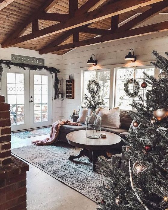 a cozy rustic living room with a round wooden table, a brick fireplace, a wooden ceiling with beams and an evergreen garland
